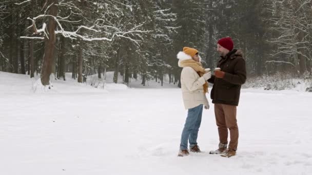 冬の森の中の白い雪の牧草地に立つ多民族のロマンチックなカップルのワイドショット ホットドリンクと紙コップを保持 その後 白人のボーイフレンドは彼のアジアのガールフレンドを抱擁 — ストック動画