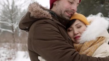 Açık havada güzel bir Asyalı kadını kucaklayan mutlu beyaz adamın orta boy yakın çekimi. Kış kıyafetleri giymiş romantik bir çift randevuya çıkıyor.
