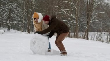 Sıcak kıyafetler giymiş, gülümseyen, kış ormanı zemininde yuvarlanan büyük bir kar topu gibi neşeli beyaz bir erkek ve Asyalı kadın. Kardan adam yapan çift
