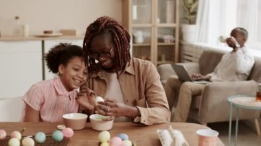 Paskalya için Afrika 'nın genç anne boyama yumurtasının orta boy fotoğrafı, kadına yaslanan ve izleyen kızı, yemek odasında masada oturan, dizüstü bilgisayarlı babası arka plandaki koltukta çay içen babası.