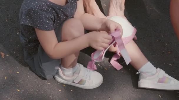 高角镜下 金发白人小女孩穿着休闲装和白色运动鞋坐在柏油路上 而她的不知名妈妈帮助她穿上膝盖垫 — 图库视频影像