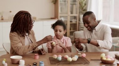 Orta ölçekli mutlu Afrikalı aile yemek masasında oturuyor, konuşuyorlar, baba, anne ve kız renkli Paskalya yumurtası değiş tokuşu yapıyorlar.