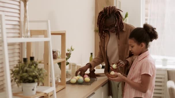 若いアフリカ人女性と台所に立つ10歳の少女の側面図 テーブルを切断から色イースターエッグをボウルに入れ 女の子歩くとそれを運びます — ストック動画