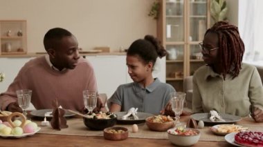 Orta ölçekli mutlu Afrikalı bir adam okul çağındaki kızıyla konuşuyor, gülümsüyor, sonra kız anne ve babasıyla el ele tutuşuyor, aile şenlikli yemek masasında oturuyor, gözlerini kapıyor ve dua ediyor.