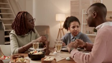 Okul çağında at kuyruklu Afrikalı bir kızın orta ölçekli yakın çekimi, festival gecesi yemek masasında oturup genç anne ve babanın canlı sohbetlerini dinlemek ve izlemek.