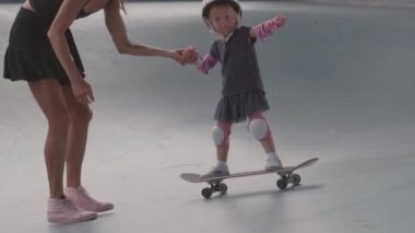 Gerçek zamanlı olarak, beyaz kadın sokak kıyafetleri giyip kızının elini tutuyor. Emniyet kaskı, diz ve dirseklik takıyor ve kaykay parkında kaykay yapmasına yardım ediyor.