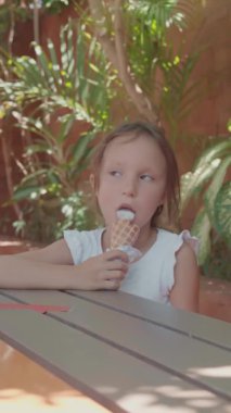Kahverengi saçlı, beyaz tişörtlü, tahta masada oturan ve dondurma külahı yiyen beyaz kızın dikey görüntüsü.