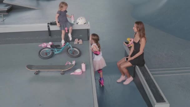 年轻漂亮女人穿着休闲装坐在室内溜冰场的斜坡上 看着两个女儿玩乐的高视角画面 — 图库视频影像