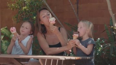 Orta boy beyaz kadın ve iki küçük kızı açık havada ahşap masada oturmuş dondurma yiyorlar.