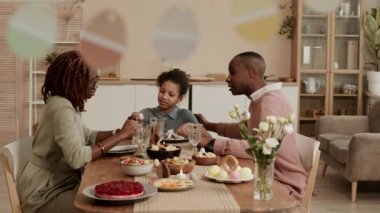 Orta ölçekli mutlu bir siyahi aile masada oturuyor, konuşuyor, şenlikli bir akşam yemeği yiyor, odada asılı duran kağıt paskalya yumurtalarının süslü çelenklerine takılıyor.