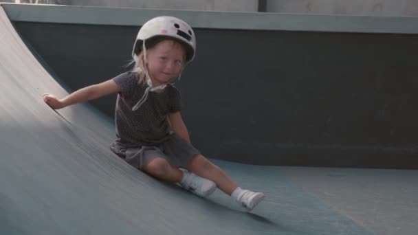 陽気な小さなブロンド白人の女の子は スカートを着て Tシャツと安全ヘルメットは 屋内スケート場で人工丘の上に登るしようとします カメラを見て 離れて実行して — ストック動画