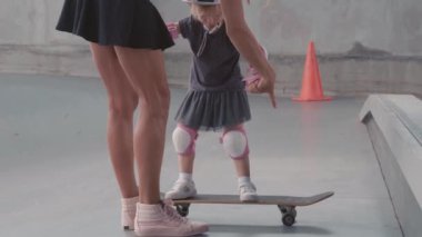 Sıska bir kadının sokak kıyafetleri içinde kızının elini tutarken renkli bir kaskla dizlik ve dirsek yastığı takarken ve ona kaykay parkında nasıl kayılacağını öğretirken.