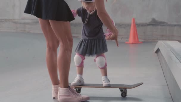 バックビューの若いですスリム女性身に着けていますストリートウェア保持手の彼女の小さな娘身に着けていますカラフルな安全ヘルメット 膝と肘パッドと彼女を教える方法スケートパークでスケートボード — ストック動画