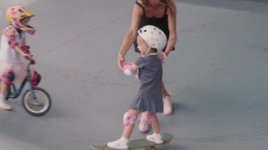 Kızının el ele tutuşup, emniyet kaskı ve pedleri taktığı ve kaykay yapmasına yardım ettiği yüksek açılı bir manzara.