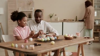 Orta uzunlukta gururlu bir Afrikalı baba konuşuyor ve neşeli kızının Paskalya yumurtalarını boyamasını izliyor, yemek masasında oturuyor, anne arka planda mutfakta yemek pişiriyor.