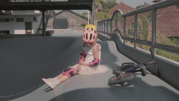10代の白人の少女の広いショット彼女の顔にヘルメットと屋外スケートパークのランプの上に座っているカジュアルな服 肘と膝パッドを身に着けているし それを脱いで — ストック動画