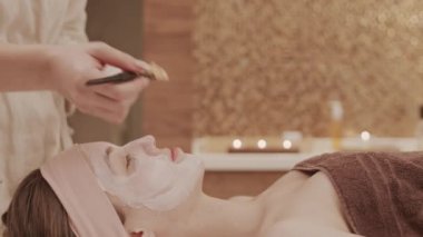 Spa merkezindeki masaj masasında uzanan beyaz bir kadın ve yüzüne kilden maske takan tanınmayan bir kozmetikçi.