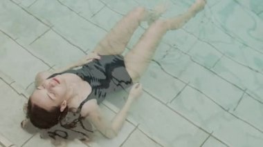 Uzun kahverengi saçlı, tek parça mayo giyen beyaz bir kadın yüzme havuzunda mermer merdivenlerde gözleri kapalı ve dinleniyor.