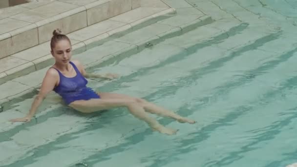 白种人的年轻女子 金发碧眼 身穿蓝色游泳衣 坐在大理石楼梯上 躺在游泳池里休息 — 图库视频影像