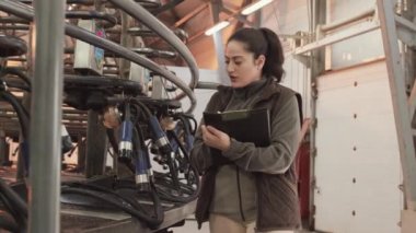 Orta uzunluktaki melez kadın işçiler ayakta duruyor, notlar alıyor, dönen endüstriyel sağma makinesinin çalışabilirliğini kontrol ediyor. 