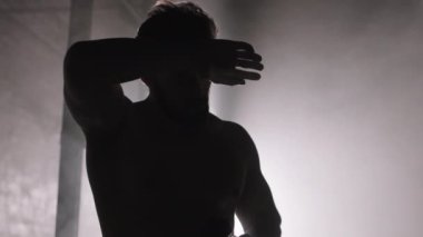 Spor salonunda karanlıkta duran kaslı bir adamın silüeti ve yoğun işlevsel eğitimden sonra spor şişesinden su içmesi.