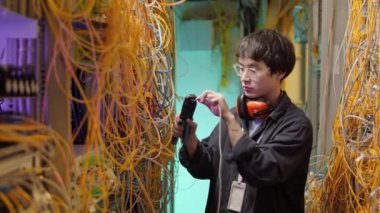 Medium shot of young Asian IT technician wearing streetwear, glasses and headphones on his neck standing in server room and connecting wires