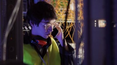 Medium shot of young Asian IT engineer wearing special uniform and glasses standing in data center, making photo of wires using smartphone camera and then sending photo to manager
