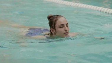 Mavi mayo giyen, kapalı havuzda kurbağalama yüzen Kafkasyalı genç bir kadın.
