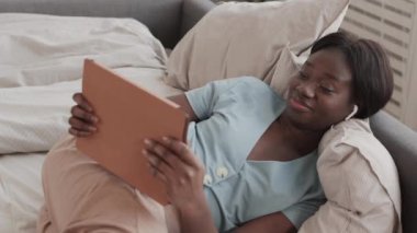 Kablosuz kulaklık takan, elinde dijital tabletle yatıp video oyunu oynayan Afro-Amerikan genç bir kadının yüksek açılı görüntüsü.