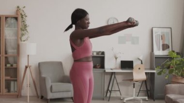 Orta boy Afro-Amerikan spor kadın, şık pembe spor giyerek oturma odasında dambıllarla egzersiz yaparken.