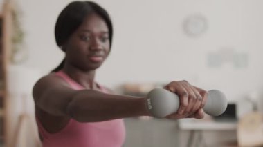 Pembe kolsuz tişört giyen genç Afro-Amerikan sporcuların kilitlenmesi. Oturma odasında otururken dambıllar kullanarak kol kasları pompalaması.