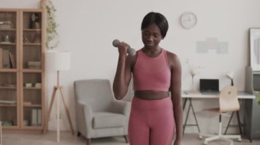 Orta boy Afro-Amerikan spor kadın spor giyerken oturma odasında dambıllarla egzersiz yaparken.