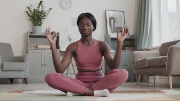 穿着粉色运动服 穿着运动鞋 头戴莲花的年轻的非洲裔美国瑜伽队员 坐在客厅里 闭着眼睛 沉思着 — 图库视频影像