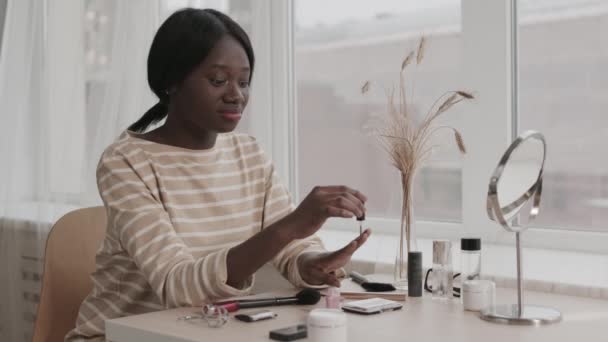 Lockdown of young lovely African-American woman wearing casual clothes sitting at table with different cosmetic products on it and applying nail polish on her nails