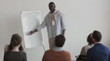 Orta uzunluktaki Afrikalı genç bir ofis çalışanı tanınmayan izleyicilerin önünde konuşuyor, beyaz tahtaya grafik çiziyor, iş sunumu yapıyor.