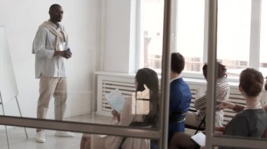 Genç Afrikalı erkek konuşmacının iş sunumu yaparken, ofiste meslektaşlarının önünde dikilirken, seyircilerle iletişim kurarken, ön planda cam kapılar kayarken.