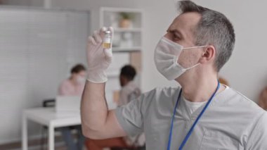 Medikal eldiven, yüz maskesi, önlük takan, hastanede dikilen, ampulü alüminyum kapakla tutan ve sonra da kameraya bakan beyaz erkek doktorun orta boy yakın çekim POV 'si.