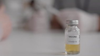 Yüzeyde metal kapaklı, sarı sıvılı ve Covid-19 aşılı sabit bir ampul görüntüsü var. Arka planda bulanık hareketler oluyor.