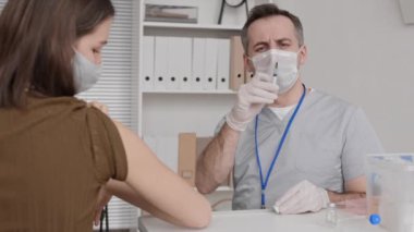 Tıbbi maske ve eldiven takan, şırınga tutan, hastanın önünde oturan, sonra da koluna grip aşısı yapan, konuşan neşeli beyaz erkek doktorun göğsü yukarıda.