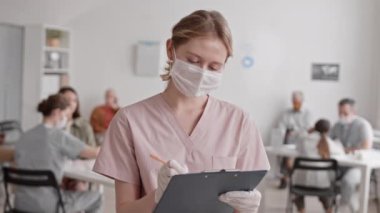Önlük, tıbbi maske ve eldiven takan, pano tutan ve üzerine yazı yazan beyaz kadın doktorların orta boy yakın çekim görüntüsü.