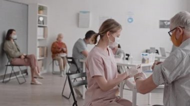Tıbbi maske ve eldiven takan orta boylu, sarışın, beyaz kadın doktor dolu bir aşı odasında oturuyor ve gümüş saçlı hastaya iğne yapıyor.