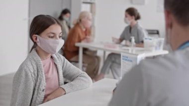 Yüksek açılı, orta ölçekli, koruyucu yüz maskesi takan, klinikte masada oturan, tanınmayan bir tıp çalışanı tarafından aşıya uğrayan, sonra da ayrılan beyaz bir kız.