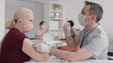 Maske takan, eldiven takan, hastanede oturan, önünde oturan yaşlı ve sarışın kadına aşı yapan beyaz bir erkek doktorun yan görüntüsü.