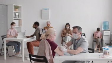 Orta uzunlukta, maske ve eldiven takan beyaz bir erkek doktor, masada oturuyor, hastane odasındaki yaşlı sarışın kadına aşı aşısı yapıyor, arka planda bekleyen hastalar...