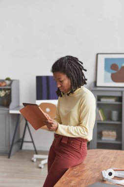 Tablet kullanan genç Afrikalı Amerikalı kadının dikey portresi. İç kısımda masa başında dururken.