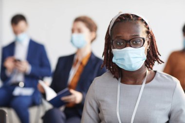 İş konferansında izleyiciler arasında otururken maske ve gözlük takan genç Afro-Amerikalı iş kadınının ön manzara portresi.