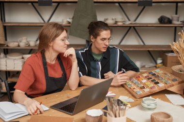 Çömlek atölyesinde dizüstü bilgisayarla ahşap masada çalışırken küçük işletmeleri yöneten iki gencin portresi.