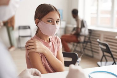 Pembe maske takan ve klinikte aşı beklerken kameraya bakan güzel bir kızın portresi.