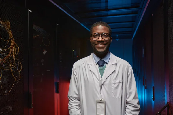 Waist up portrait of African American data scientist wearing lab coat and smiling at camera while working with supercomputer in server room, copy space