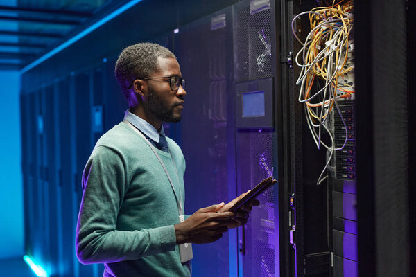 Side view portrait of African American data engineer holding digital tablet while working with supercomputer in server room lit by blue light, copy space